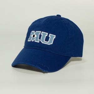 Puede incluir: Gorra de béisbol azul marino con las letras "MU" bordadas en la parte delantera. La "M" presenta un diseño de ojo. La gorra tiene una visera curva y un aspecto desgastado.