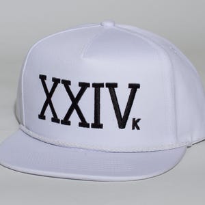 Pode incluir: Um boné snapback branco com os algarismos romanos XXIV bordados em fio preto no painel frontal. O chapéu inclui um detalhe de corda branca a condizer na base da coroa e uma aba plana, proporcionando um visual limpo e estruturado.