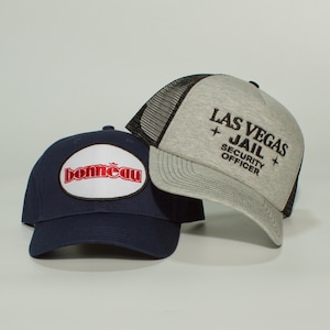 Könnte beinhalten: Zwei Baseballkappen. Eine ist marineblau mit einem weißen und roten ovalen Aufnäher mit der Aufschrift "bonneau". Die andere ist grau mit einem schwarzen Mesh-Rücken und dem Text "LAS VEGAS + JAIL + SECURITY OFFICER".