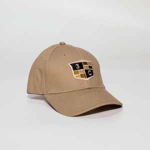 Può includere: Cappellino da baseball beige con visiera curva. Il cappellino presenta un emblema ricamato con il numero "3" e la lettera "C" su un disegno a scudo. Il cappellino è realizzato in tessuto resistente e ha una vestibilità strutturata.