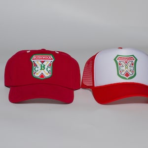 May include: Two baseball caps featuring a shield-shaped patch with the text BUSHWOOD and CBC. The left cap is solid red, while the right cap has a white front panel, a red brim, and red mesh back panels. Both caps display the same embroidered emblem.