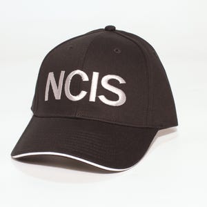 May include: Black baseball cap with the embroidered text "NCIS" in silver. The cap has a curved brim with a white trim. The hat is a classic six-panel design.