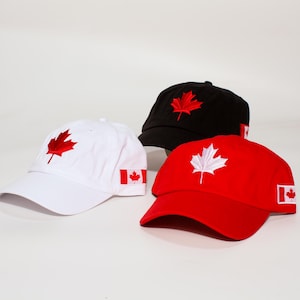 Op de afbeelding: Drie baseballpetten met de Canadese vlag. Een witte pet met een rode esdoornblad, een zwarte pet met een rood esdoornblad en een rode pet met een wit esdoornblad. Elke pet heeft de Canadese vlag op de zijkant.