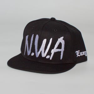 Puede incluir: Gorra de béisbol negra con el texto bordado "N.W.A" en una fuente estilizada en la parte delantera. La palabra "Eazy" está bordada en el lateral. La gorra tiene una visera plana.