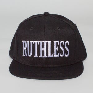 Puede incluir: Gorra de béisbol negra con la palabra "RUTHLESS" bordada en blanco en la parte delantera. La gorra tiene una visera plana y una corona redondeada. El sombrero está hecho de una tela texturizada.
