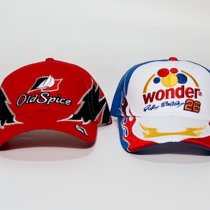 Peut inclure: Deux casquettes de baseball. L'une est rouge avec "Old Spice" et un logo. L'autre est blanche avec un bord bleu, le mot "wonder" et le numéro "26". Les deux ont des visières incurvées et sont brodées.