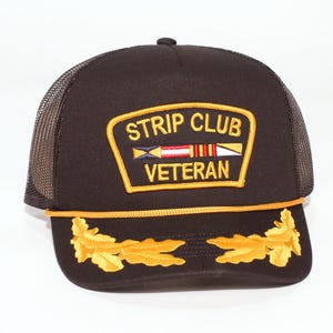 May include: A black trucker hat with a mesh back and a curved brim. The front of the hat features a patch with the words "STRIP CLUB VETERAN" in gold lettering. Gold leaf designs adorn the brim.