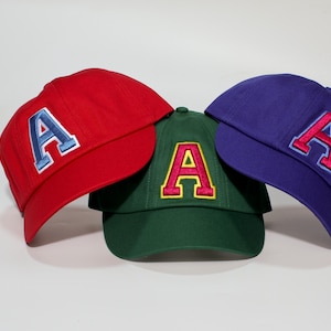 May include: Three baseball caps in red, green, and purple. Each cap features a large embroidered letter "A" in contrasting colours. The red cap has a blue and white "A", the green cap has a red and yellow "A", and the purple cap has a red and white "A".