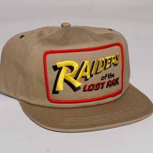 May include: A tan baseball cap with a flat brim. The cap features a rectangular patch with the words "Raiders of the Lost Ark" embroidered in yellow and black, outlined in red. The cap has a button on top.