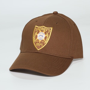 May include: A brown baseball cap with the King County Sheriff's Dept. emblem. The emblem is a shield shape with gold and white detailing, and the text "SHERIFF'S DEPT." and "SINCE 1816" are visible.