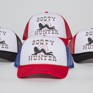 May include: Four trucker hats with white fronts, mesh backs, and curved brims. Each hat features the text "BOOTY HUNTER" above a silhouette of a woman in a crosshair target. The brims and mesh backs are in black, red, and blue.
