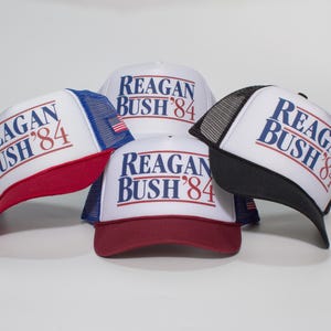 Op de afbeelding: Vier witte truckerpetten met de tekst "REAGAN BUSH '84" in blauw en rood. Elke pet heeft een verschillend gekleurde rand en mesh-achterkant: rood, blauw, zwart en wit. De petten worden op een witte achtergrond weergegeven.