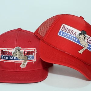 May include: Two red baseball caps with the "Bubba Gump Shrimp Co." logo. One cap is a classic style, and the other is a trucker hat with a mesh back. The logo features a cartoon shrimp.
