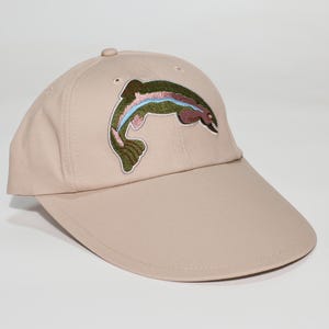May include: A beige baseball cap with an embroidered rainbow trout. The fish design incorporates green, blue, pink, and brown hues. The cap has a curved peak and a button on the crown. A practical accessory for fishing or outdoor activities.