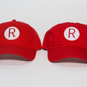 May include: Two red baseball caps with a white circle and a capital "R" embroidered on the front. The caps have a curved brim and a rounded crown. The caps are displayed on a white background.