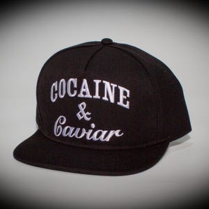 以下が含まれることがあります： 黒いベースボールキャップ。前面に白で「COCAINE & Caviar」の文字が刺繍されています。つばは平らで、クラウンは丸みを帯びています。クラシックなスタイルです。