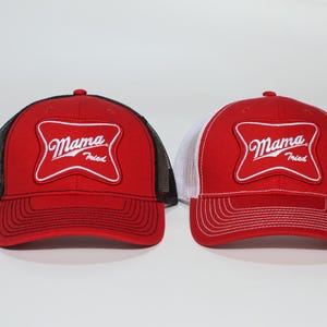 May include: Two red trucker hats with the words "Mama Fried" in a white, stylized font inside a white outline. One hat has a black mesh back, and the other has a white mesh back. Both have a red brim.