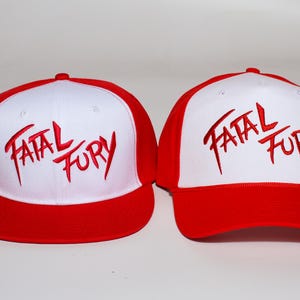 Op de afbeelding: Twee rode en witte baseballpetten. De petten hebben een rode klep en een wit voorpaneel met de geborduurde tekst "FATAL FURY" in het rood. De petten worden naast elkaar weergegeven tegen een witte achtergrond.