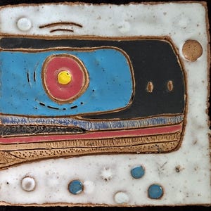 May include: Square ceramic tile with a retro cassette tape design. The tile features a blue cassette body with a red and yellow circle, black details, and a white border with blue and brown accents. The tile is approximately 6 inches square.