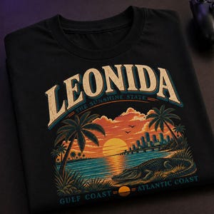 May include: A black t-shirt featuring a graphic print with the text LEONIDA, THE SUNSHINE STATE, GULF COAST, and ATLANTIC COAST. The design depicts a sunset over water with palm trees, a city skyline, and an alligator resting on the shore.