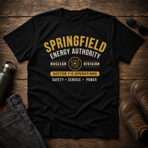 May include: A black short-sleeve t-shirt featuring a gold and cream graphic that reads Springfield Energy Authority, Nuclear Division, Sector 7-G Operations, Safety, Service, Power, with a central atom symbol icon printed on the chest.