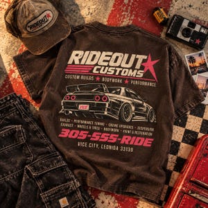 May include: A dark brown t-shirt featuring a graphic of a sports car and the text RIDEOUT CUSTOMS, CUSTOM BUILDS, BODYWORK, PERFORMANCE, 305-555-RIDE, and VICE CITY, LEONIDA 33139. The design includes a red star and pink text accents.