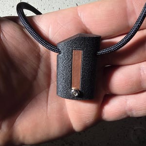 Può includere: Una collana con pendente nero dal design geometrico. Il pendente ha una forma rettangolare con un accento color rame e una piccola gemma trasparente. Il cordino della collana è nero e intrecciato.