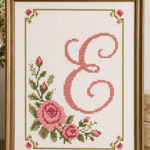 Può includere: Opera d'arte a punto croce incorniciata con la lettera "E" rosa circondata da rose rosa e foglie verdi. L'opera è incorniciata in una cornice di legno color oro con un bordo dorato e accenti floreali agli angoli. Lo sfondo è bianco.