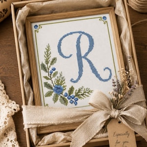 May include: A framed cross-stitch embroidery featuring a large blue letter R surrounded by green foliage and blue berries. The piece is presented in a cardboard gift box lined with tissue paper, tied with a beige ribbon and a tag reading Especially for you.