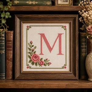 May include: A framed cross-stitch with the letter "M" in pink, encircled by a floral design of pink roses and green foliage. The dark brown wooden frame sits on a shelf with antique books. The piece is a decorative item.