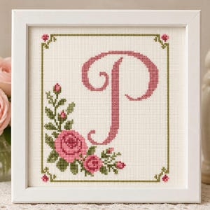May include: A framed cross-stitch featuring the letter "P" in pink, surrounded by a floral arrangement of pink roses and green leaves. The design is set against a cream-colored background, framed in a white square.