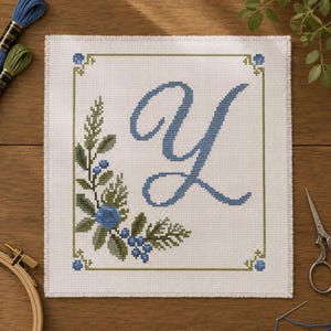 May include: A square white fabric piece featuring a cross-stitch design of the letter Y in blue. The letter is surrounded by a green and blue botanical border with leaves and berries. The edges of the fabric are finished with a stitched border.