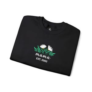 Pode incluir: Camisola preta com um design floral com duas margaridas brancas com centros amarelos e folhas verdes. A palavra "mama" é bordada sob as flores, com "EST. 2002" por baixo. A camisola está dobrada cuidadosamente sobre uma superfície branca.