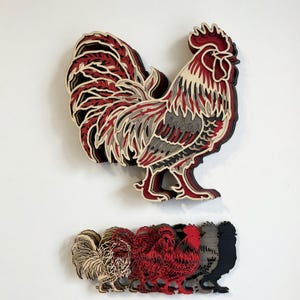May include: A layered wooden rooster wall art piece in red, black, gray, and cream colors. Below, a row of smaller rooster cutouts in various colors are displayed. The rooster has a detailed design, with layered feathers and a prominent comb.