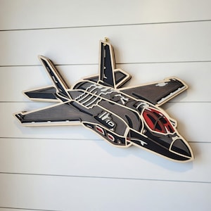 May include: A multi-layered wooden F-35 fighter jet wall art piece. The artwork features a detailed design with grey, black, white, and red accents. The jet is mounted on a white wall, showcasing its intricate construction and colour palette.