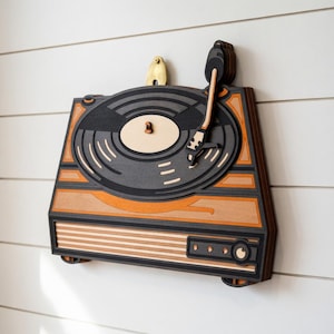 Pode incluir: Arte de parede em madeira retratando uma vitrola vintage. A arte apresenta um design em camadas com tons de preto, cinza, laranja e madeira natural. A vitrola tem um toca-discos, braço e botões de controle.