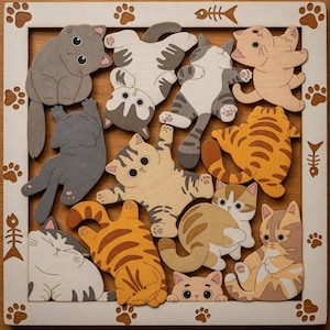 Pode incluir: Um quebra-cabeça de madeira com formas de gatos em tons de cinza, bege e laranja. As peças estão dispostas dentro de uma moldura branca decorada com pegadas e motivos de espinha de peixe. Os gatos são retratados em poses brincalhonas.