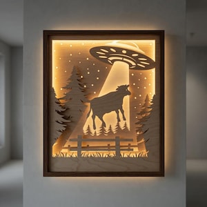 May include: Wooden framed art piece depicting a cow being abducted by a UFO. The artwork features a layered design with a warm, glowing light illuminating the scene. Silhouetted trees, a fence, and a starry sky complete the rural landscape.
