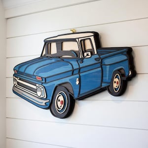 May include: A blue wooden pickup truck wall art piece. The truck has a white roof, black tyres, and a detailed design. The artwork is mounted on a white shiplap wall, creating a rustic aesthetic.