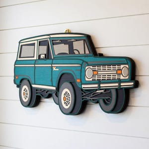 May include: A teal and white wooden wall hanging of a vintage Bronco SUV. The layered design features a white roof, silver accents, and orange details on the front grille. The wheels are gray and white.