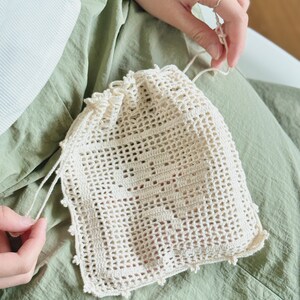 May include: A cream-colored drawstring pouch crafted from crochet with an openwork mesh pattern. The front features a central square motif with a subtle, integrated geometric design. The top edge is gathered with matching crochet strings.