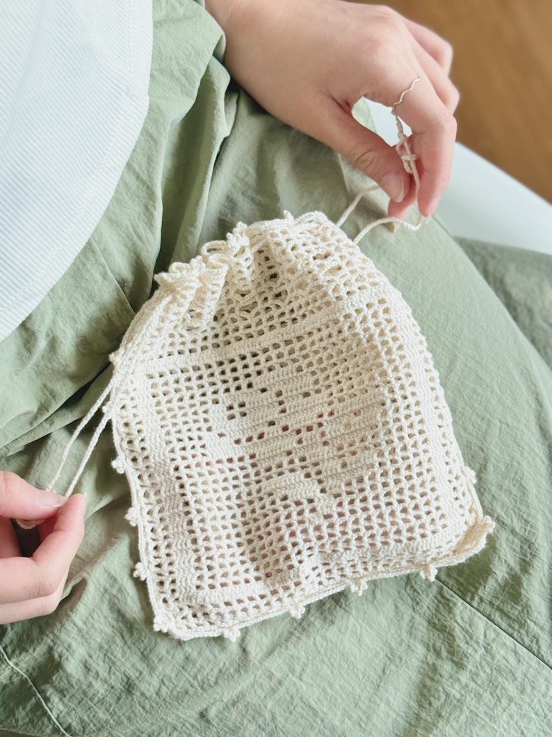 May include: A cream-colored drawstring pouch crafted from crochet with an openwork mesh pattern. The front features a central square motif with a subtle, integrated geometric design. The top edge is gathered with matching crochet strings.