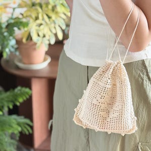May include: A cream-colored drawstring pouch crafted with an intricate openwork crochet pattern. The bag features a square base, a grid-like mesh body, and thin cord handles that cinch the top closed, creating a gathered effect at the opening.