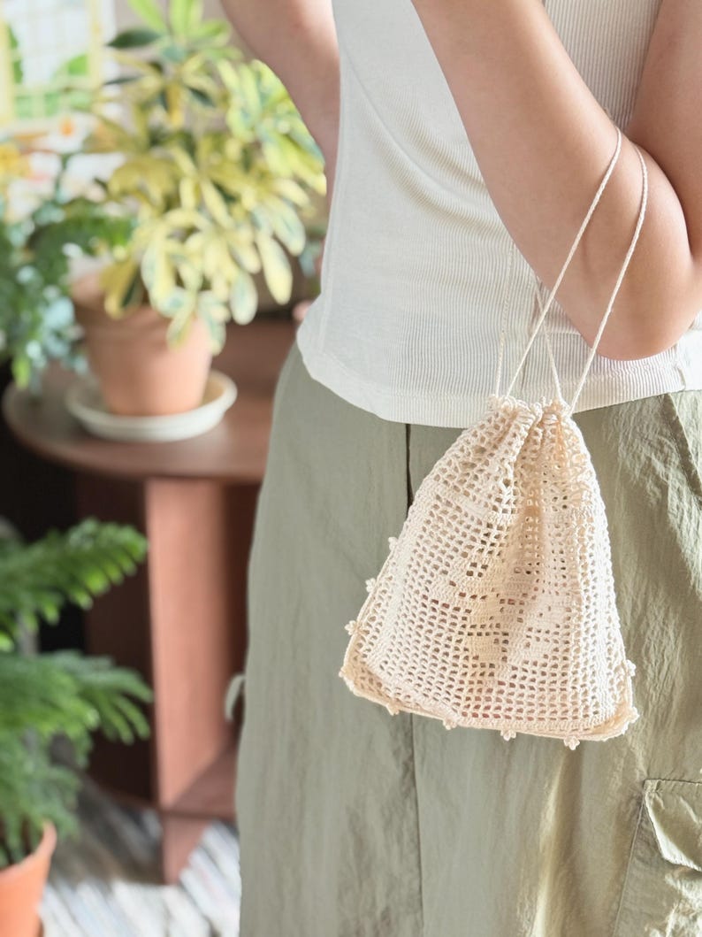 May include: A cream-colored drawstring pouch crafted with an intricate openwork crochet pattern. The bag features a square base, a grid-like mesh body, and thin cord handles that cinch the top closed, creating a gathered effect at the opening.