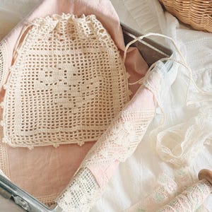 May include: A cream-colored crocheted drawstring pouch featuring a grid pattern and a scalloped edge. The pouch rests on a pale pink fabric inside a metal container, accompanied by a spool of floral lace trim and additional white lace fabric.