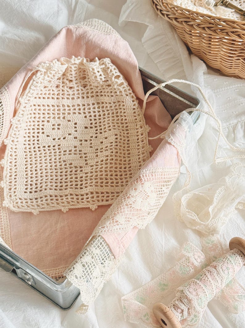 May include: A cream-colored crocheted drawstring pouch featuring a grid pattern and a scalloped edge. The pouch rests on a pale pink fabric inside a metal container, accompanied by a spool of floral lace trim and additional white lace fabric.