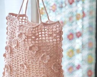 May include: A light pink crochet tote bag with flower appliqués. The bag has a rectangular shape with a scalloped edge and long handles. The bag is hanging from a white hook, with a floral patterned curtain in the background.