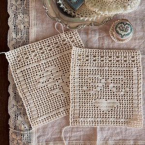 May include: Two square off-white crochet doilies featuring a central heart pattern, displayed on a light beige linen tablecloth with a wide lace border. A silver tray holds additional crochet items, a small blue box with a stone, and a round tin.