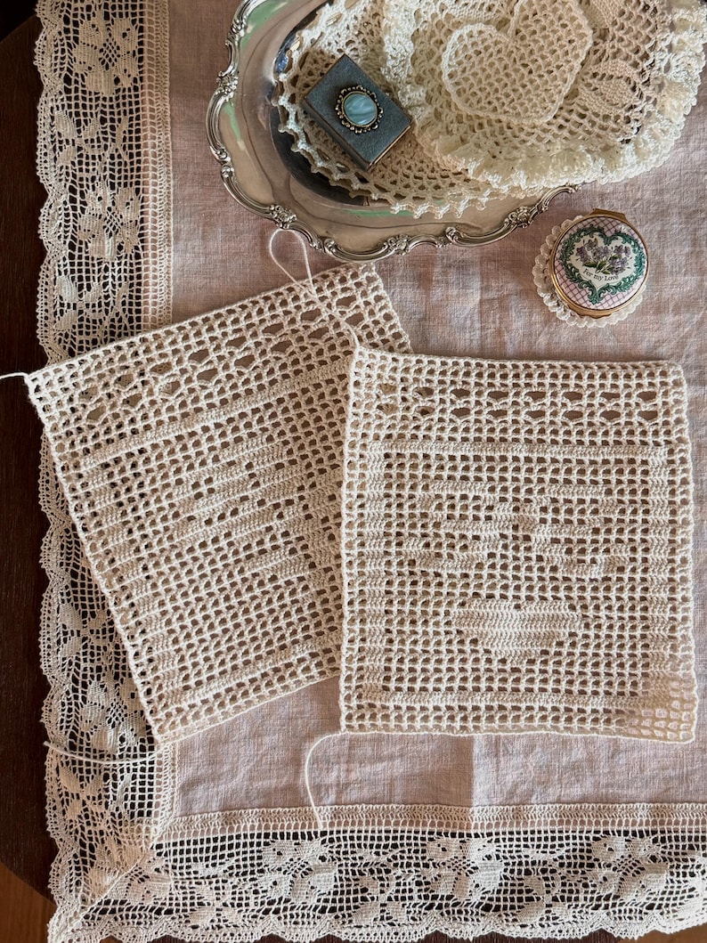 May include: Two square off-white crochet doilies featuring a central heart pattern, displayed on a light beige linen tablecloth with a wide lace border. A silver tray holds additional crochet items, a small blue box with a stone, and a round tin.