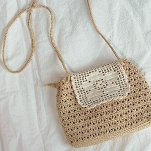 May include: A beige crochet crossbody bag featuring an open-weave pattern and a long, thin shoulder strap. The bag includes a rectangular flap made of white crochet with a scalloped edge, contrasting against the tan body of the bag.